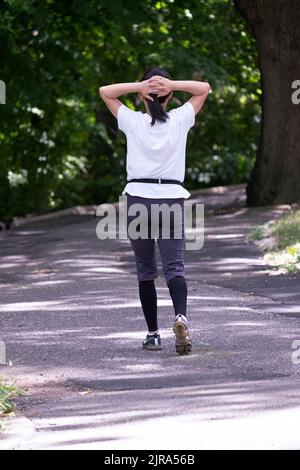 Una donna asiatica americana non identificata multitasking da camminare esercizio e stretching simultaneamente. In un parco a Queens, New York. Foto Stock