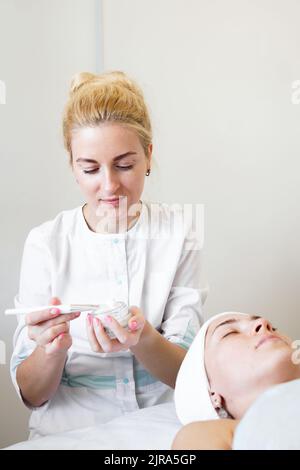 Il cosmetologist che fa la procedura anti-età applica l'acido di peeling, giovane cliente attraente femminile alla clinica di bellezza. Terapia sanitaria Foto Stock