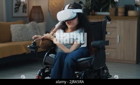 Donna felice con una disabilità in una sedia a rotelle elettrica che guida in tutta la stanza a casa in cuffia per realtà virtuale utilizzando il metaverse Foto Stock