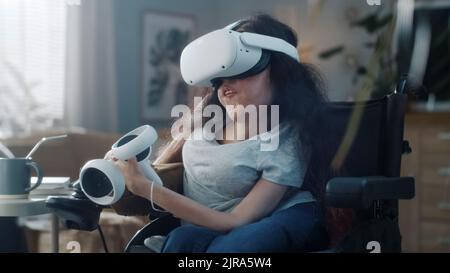 A casa donna con disabilità in sedia a rotelle indossando cuffie per realtà virtuale con controller si collega al metaverse e si guarda intorno Foto Stock