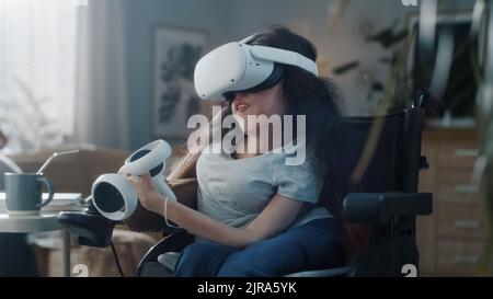 A casa donna con disabilità in sedia a rotelle indossando cuffie per realtà virtuale con controller si collega al metaverse e si guarda intorno Foto Stock