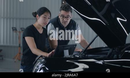 Uomo e donna dipendenti vicino al cofano che eseguono la diagnosi e il controllo di un'auto insieme in un'officina di riparazione, utilizzando un tablet e discutendo della riparazione Foto Stock