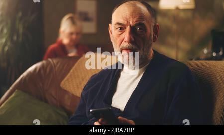 L'uomo anziano depresso sul divano togliendo gli occhiali mentre tiene il telefono con le notizie difettose e preoccupato Foto Stock