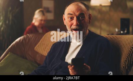 Sorpreso uomo anziano togliendo gli occhiali e guardando la macchina fotografica in stupore, dopo aver letto buone notizie sul cellulare mentre si siede sul divano in soggiorno a casa Foto Stock