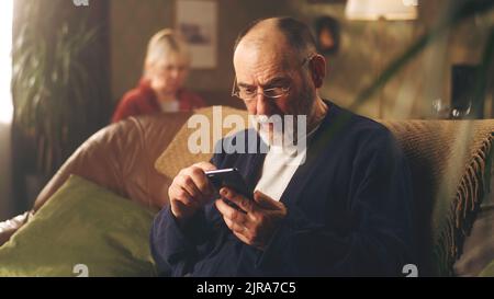 L'uomo anziano depresso sul divano togliendo gli occhiali mentre tiene il telefono con le notizie difettose e preoccupato Foto Stock