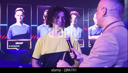 Giovane sportivo cyber sorridente e parlando con un presentatore maschio adulto mentre si trova sotto l'illuminazione al neon durante il torneo di gioco Foto Stock