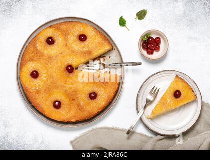 Vista dall'alto della torta rovesciata di ananas fatta in casa con ciliegie candite e un pezzo tagliato su un piatto. Dessert tropicale estivo. Foto Stock