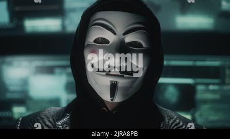 Hacker web maschile nel cappuccio e maschera anonima guardando la telecamera durante l'hacking di dati e server privato in un nascondiglio oscurato Foto Stock