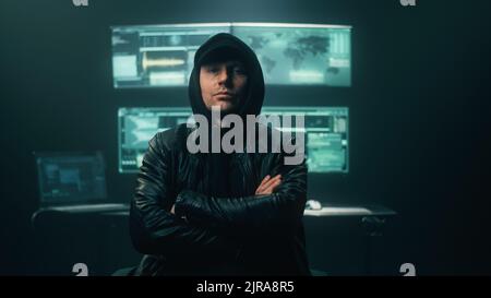 Uomo cybercriminale in cappa guardando la telecamera di fronte al tavolo con computer di pirateria informatica in camera oscura Foto Stock
