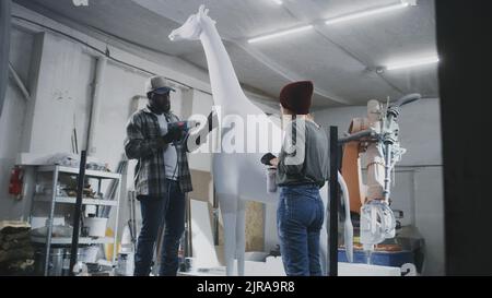 Donna che mescola la vernice in tazza e controlla la statua mentre l'uomo nero che asciuga il pigmento bianco sulla giraffa vicino alla macchina robotica di fresatura del braccio durante il lavoro Foto Stock