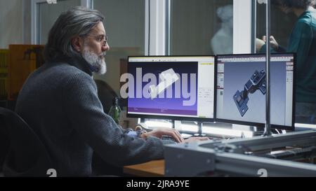 Uomo di mezza età in maglione e occhiali creando modelli 3D di dettagli sui computer mentre si siede al tavolo e si lavora in un workshop di design Foto Stock