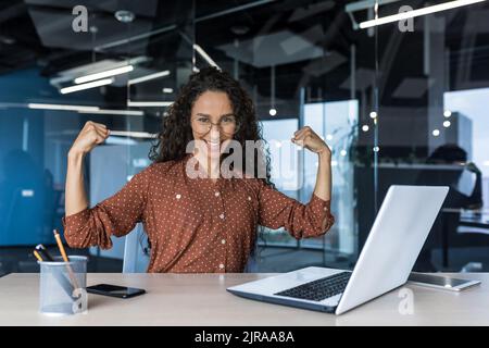 Ritratto di una donna d'affari forte che lavora all'interno di un edificio ufficio, seduto su una scrivania e utilizzando un computer portatile, sorridendo e guardando la fotocamera tenendo le mani in su gestuale superpotenza e successo Foto Stock