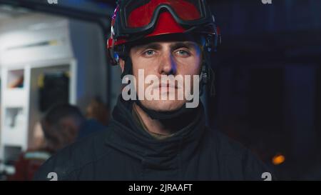 Uomo in elmetto che guarda la macchina fotografica mentre si trova vicino a un furgone ambulanza con paramedici e sopravvissuti dopo un'emergenza di notte Foto Stock