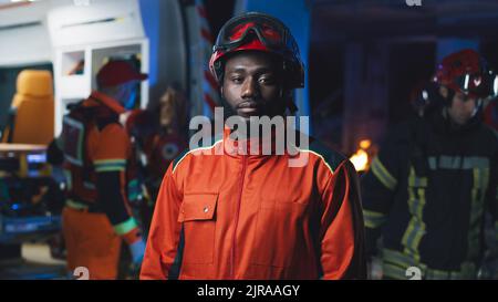 Uomo afro-americano barbuto in elmetto guardando la macchina fotografica mentre si trova vicino a furgone e colleghi durante l'incidente di notte Foto Stock