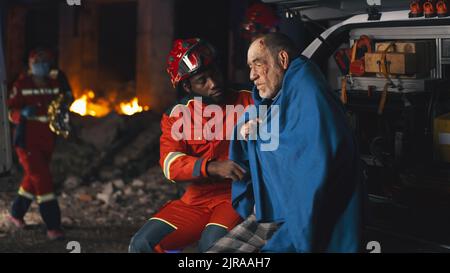 Uomo afroamericano barbuto in uniforme paramedica facendo domande all'uomo anziano avvolto in coperta dopo incidente di notte Foto Stock