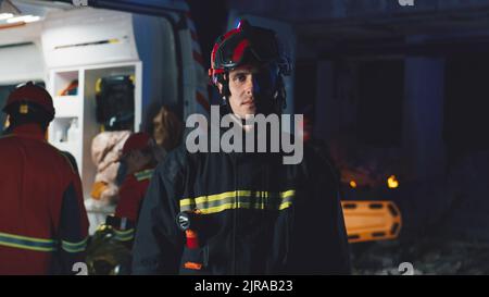 Operaio del servizio di emergenza in uniforme e hardHat che guarda la macchina fotografica mentre si trova vicino al furgone dell'ambulanza e al team di paramedici sul luogo del disastro in serata Foto Stock