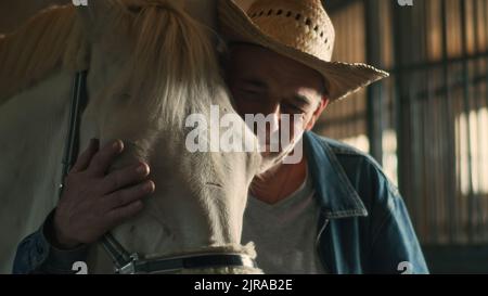 Uomo anziano positivo in cappello di paglia che abbraccia e bacia la testa di cavallo albino mentre lavora in stalla su ranch Foto Stock