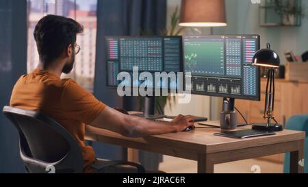 Broker indiano bearded intelligente seduto a tavola e utilizzando il computer per commerciare sul mercato azionario durante il lavoro di sera a casa Foto Stock