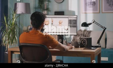 Vista posteriore di un uomo anonimo che crea un modello 3D di personaggio videoludico seduto al tavolo e lavorando a un progetto remoto a casa Foto Stock