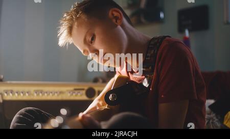 Un musicista teenager ha messo a fuoco suonando la chitarra elettrica e regolando i comandi sull'amplificatore mentre si siede a letto durante la sessione musicale nella stanza da letto illuminata dal sole a casa Foto Stock