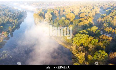 Vista aerea del drone che sorvola il fiume con la superficie d'acqua riflettente e la nebbia bianca e gli alberi della foresta sulle rive del fiume in una mattina di primo autunno soleggiato. Splendido sfondo naturale. Foto Stock