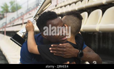 Uomo afroamericano che dà la tazza e abbraccia il ragazzo di corsa mista mentre si siede sullo stadio dopo la vittoria nella partita Foto Stock