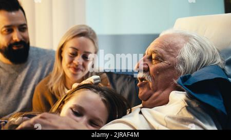 L'uomo anziano abbraccia i bambini felici vicino la madre ed il padre adulti che visitano il paziente nel reparto dell'ospedale Foto Stock