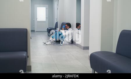 Un medico di mezza età stanco che tira fuori la protezione del viso e tocca la testa mentre si siede sul pavimento e si rilassa durante un turno difficile in clinica contemporanea Foto Stock
