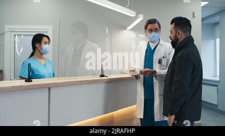 Donna asiatica in maschera parlando con paziente e annuendo poi dando carta al medico maturo e in attesa che gli uomini di lasciare durante il lavoro Foto Stock