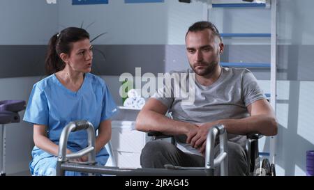 Vista del terapeuta femminile in uniforme medica che mostra la struttura a piedi e spiegare la procedura di riabilitazione a un uomo triste su sedia a rotelle in clinica moderna Foto Stock