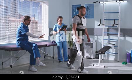 Paziente maschio adulto in exoscheletro esercitandosi sul tapis roulant mentre imparando a camminare vicino ai terapisti di riabilitazione che danno i consigli in clinica futuristica Foto Stock