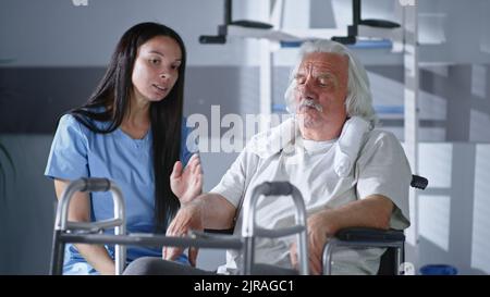 Uniforme medica che mostra la struttura a piedi e parla con l'uomo anziano su sedia a rotelle prima della sessione di riabilitazione in ospedale moderno Foto Stock