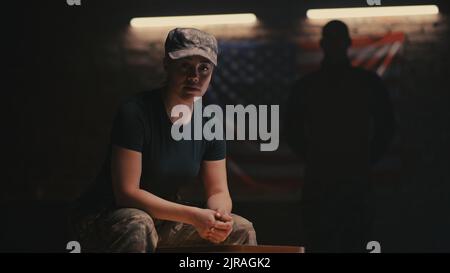Donna in mimetizzazione seduta su panca e guardando la telecamera contro la bandiera degli Stati Uniti e la silhouette del comandante su base militare Foto Stock