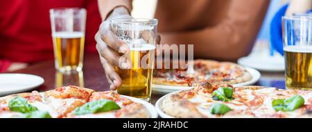 Banner orizzontale o intestazione con primo piano della mano di un ragazzo africano che prende un bicchiere di birra al ristorante pizza - Multi Ethnic Friends having fun eating pizz Foto Stock