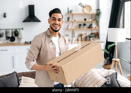 Allegro moderno indiano o arabo ragazzo, stand a casa nel soggiorno, tenendo una grande scatola di cartone, ricevuto pacco dal negozio online, preparandosi a disimballare, guarda la macchina fotografica, sorriso, emozioni felici Foto Stock