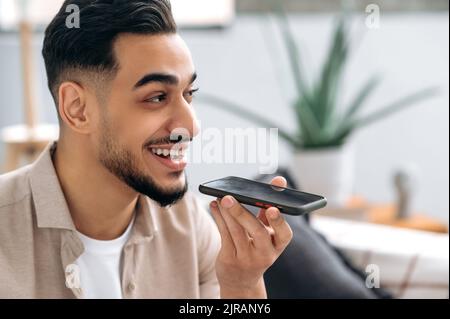 Conversazione in vivavoce. Primo piano di un eccitato uomo indiano o arabo positivo, seduto su un divano in salotto, tenendo lo smartphone, parlando in vivavoce e sorridendo, registra il messaggio vocale Foto Stock