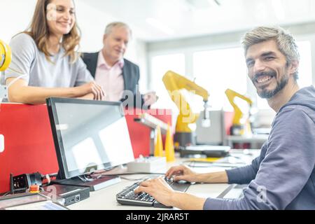 Colleghi felici che guardano un uomo sorridente che lavora al PC in una fabbrica robotica Foto Stock