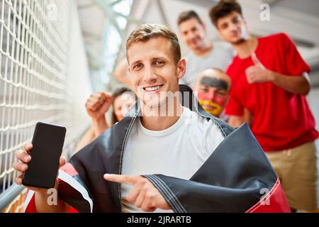 Uomo sorridente con la bandiera tedesca che indica lo schermo dello smartphone all'evento sportivo nello stadio Foto Stock