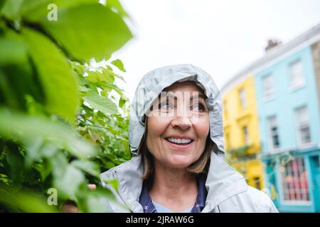 Donna anziana sorridente che indossa il rompigetto a vento dalle piante Foto Stock