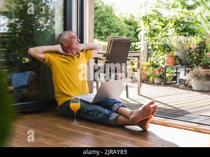 Uomo anziano sorridente con le mani dietro la testa seduto a casa Foto Stock