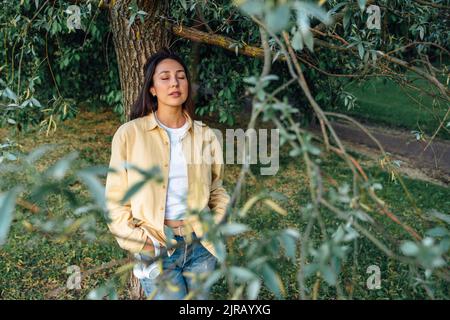 Donna con gli occhi chiusi appoggiata sull'albero Foto Stock