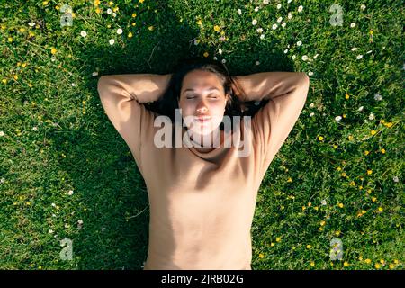 Giovane donna con le mani dietro la testa sdraiata sull'erba nel parco Foto Stock