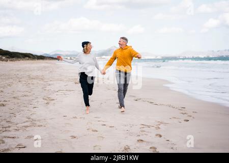 Felice coppia matura che tiene le mani e corre in spiaggia Foto Stock