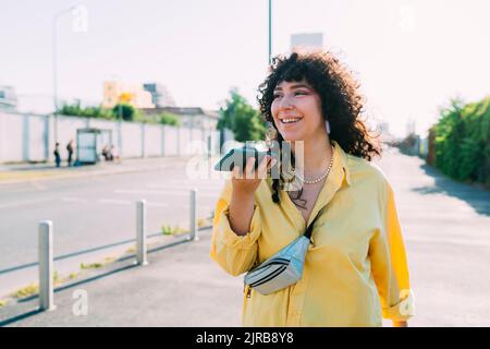 Donna sorridente che invia messaggi vocali tramite smartphone per strada Foto Stock
