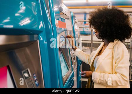 Donna con acconciatura afro utilizzando il distributore automatico di biglietti alla stazione della metropolitana Foto Stock