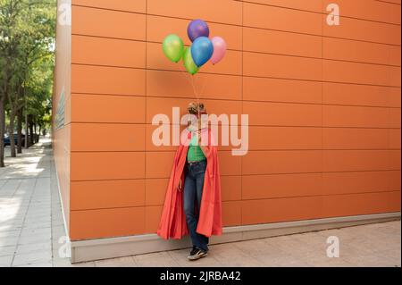 Giovane donna che indossa una maschera di coccodrillo e un mantello in piedi con palloncini multicolore davanti al muro Foto Stock