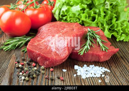 Bistecca di manzo crudo sul tagliere con verdure e spezie su sfondo di legno marrone. Bistecca di manzo, pomodoro, insalata, aglio, rosmarino, spezie. Foto Stock