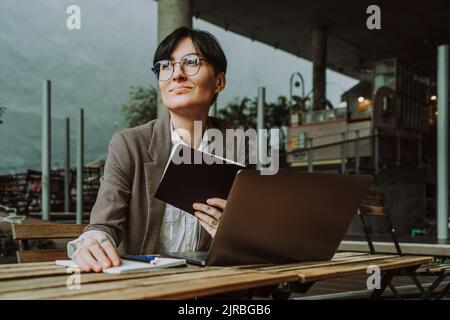 Donna d'affari premurosa e sorridente che indossa occhiali seduti con un diario e un computer portatile al bar sul marciapiede Foto Stock