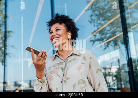 Felice donna d'affari che invia la segreteria telefonica attraverso lo smartphone di fronte alla parete di vetro Foto Stock