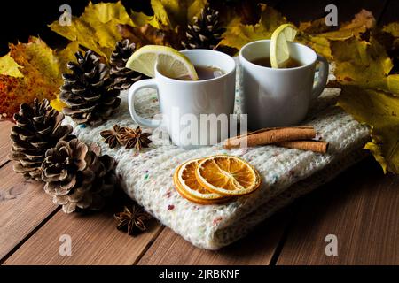 Due tazze di tè al limone su una sciarpa invernale calda a maglia. Composizione autunnale su tavolo di legno, con cannella, anice stellato, coni e foglie. Foto Stock